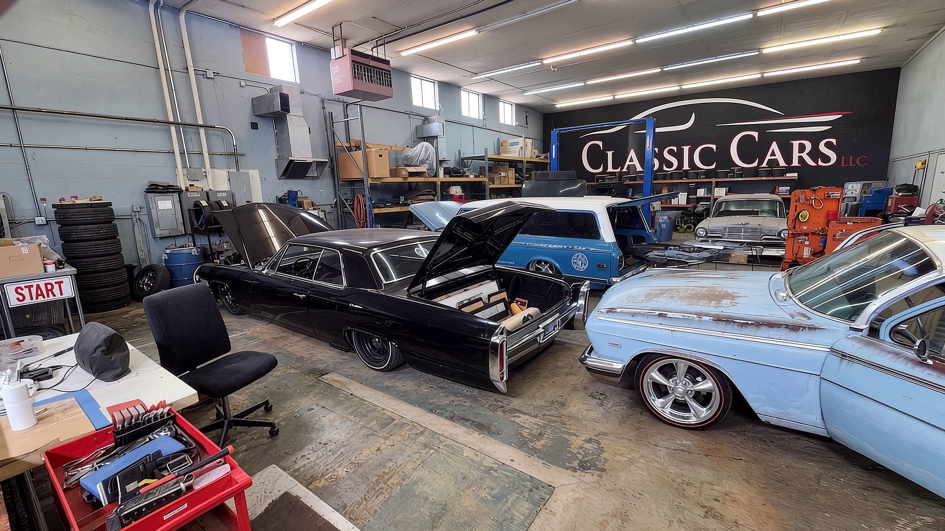 Inside the Classic Cars LLC shop: the Classic Cars logo painted across the back wall, a lowered black Cadillac with its upholstered trunk open, a patina station wagon and more classics in for work