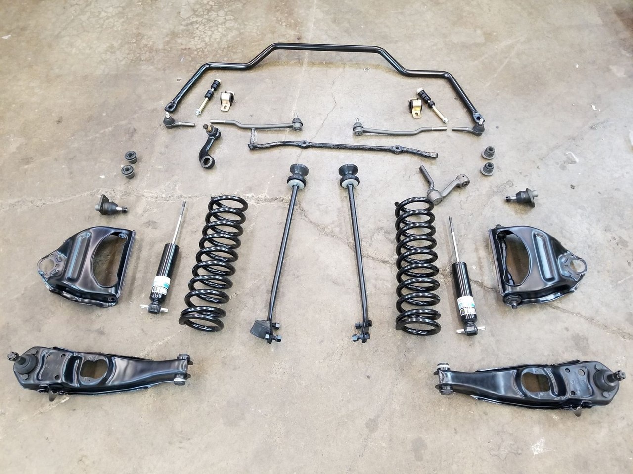 Front suspension and steering parts laid out on the shop floor: sway bar, steering linkage, coil springs, shocks, strut rods and upper and lower control arms